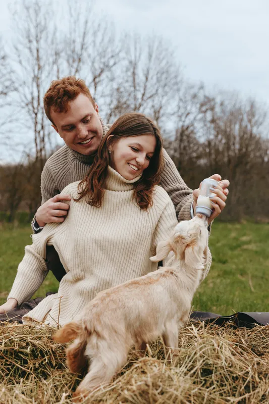 Cheerful couple outdoors, smiling with their dog, feeding moment on a hay bale, cozy sweaters, natural light, rustic field backdrop, relaxed lifestyle scene, warm tones
