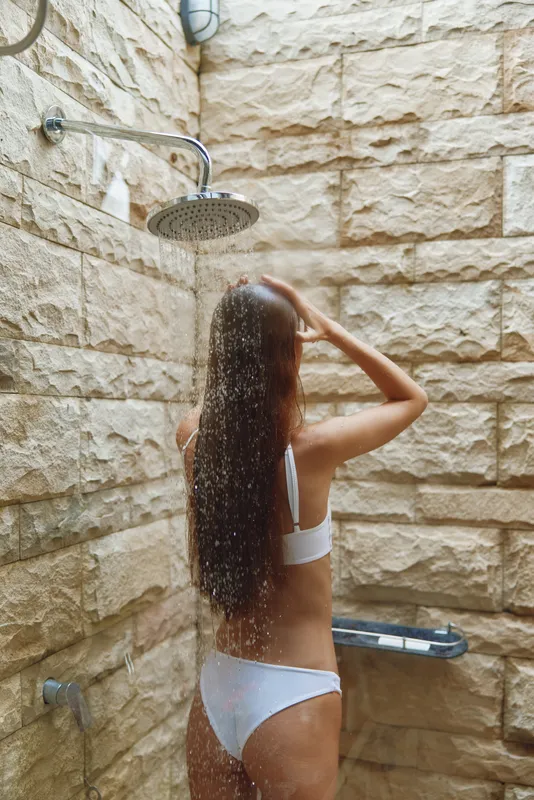 woman showering outdoors in white bikini, long hair falling down back, stone wall background, natural light and refreshing atmosphere under rainfall