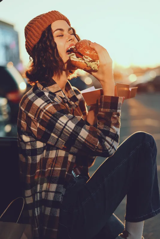 Woman eating burger outdoor in urban street at golden hour, candid lifestyle portrait showing authenticity and emotional storytelling, mindful living and warm golden hour glow.