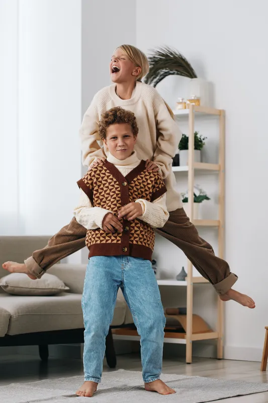 Two children play indoors, with one perched on the other's shoulders, laughing and smiling in a bright, cozy living room. A warm, playful moment.