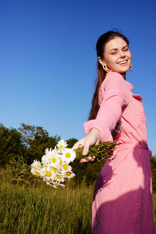 Young woman in a pink dress smiling and holding daisies against a blue sky, representing joy and nature in a vibrant summer setting