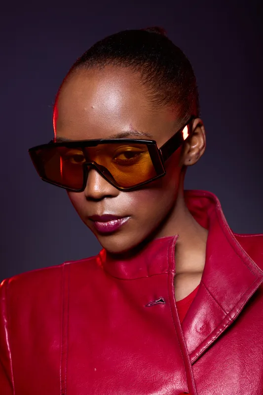 Close-up portrait of a confident black woman with dark skin wearing oversized orange sunglasses and a red leather jacket. Studio fashion concept with dramatic lighting and bold style.