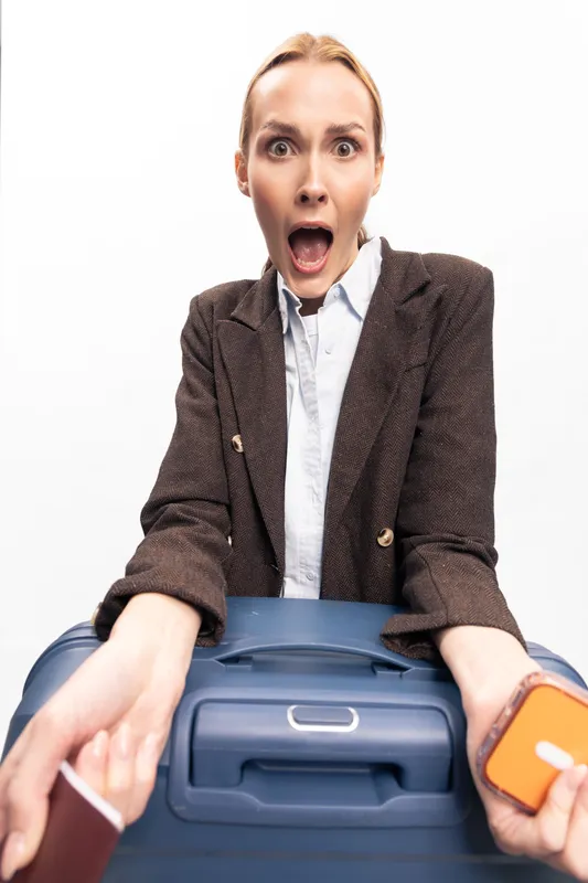 Shocked woman holding smartphone and suitcase looks surprised with wide open mouth. Travel concept, last minute packing, stress and urgency before trip. Isolated on white background.