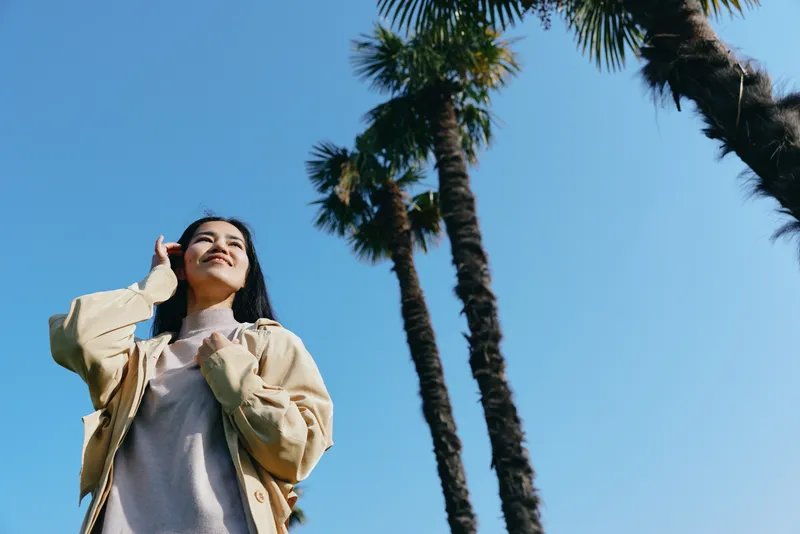 Casual outdoor scene with palm trees and a clear blue sky, a woman looking up with a thoughtful expression, wearing a light jacket and sweater, sunny day mood, relaxed travel vibe, natural light,