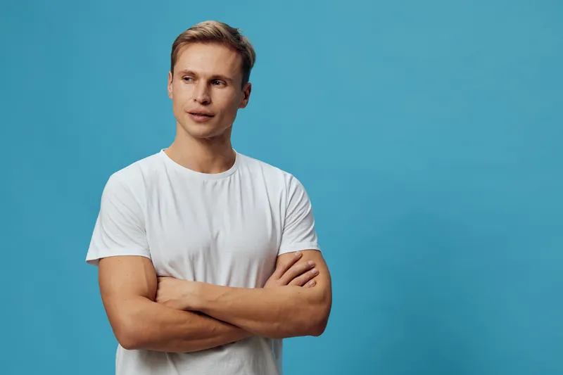 Confident young man with short brown hair wearing plain white t-shirt pose with arms crossed looking away isolated on blue background studio portrait. People lifestyle concept