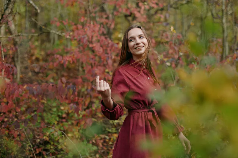 An authentic portrait features a woman in a burgundy dress in an autumn forest, smiling softly as warm light filters through colorful leaves, emphasizing natural presence.