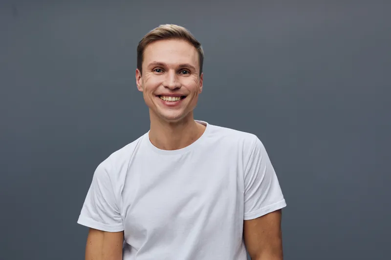 Smiling young man with short brown hair in casual white t-shirt against plain grey background, looking at camera, happy expression, indoor studio portrait, lifestyle concept, male emotion