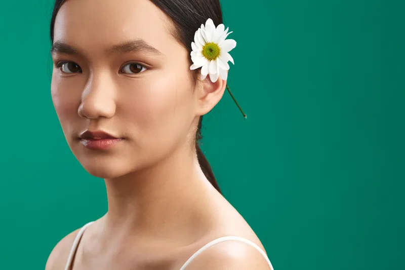 Asian woman with a flower in her hair gazes softly at the camera against a green backdrop, presenting a calm and graceful portrait that emphasizes natural beauty and serene style.