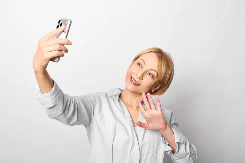 Smiling blonde woman taking a selfie with a smartphone on a clean white background, posing with one hand near face for social media communication, casual portrait concept