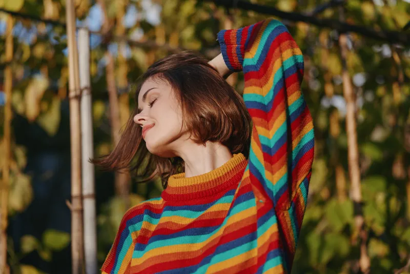 Woman portrait in colorful striped sweater smiling with eyes closed outdoors in warm sunlight, short brown hair, peaceful expression and autumn foliage background lifestyle.