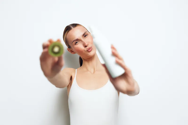 Young woman with clear skin holding kiwi slice and white bottle, skincare concept, fresh natural beauty, clean background, healthy lifestyle, cosmetic product focus