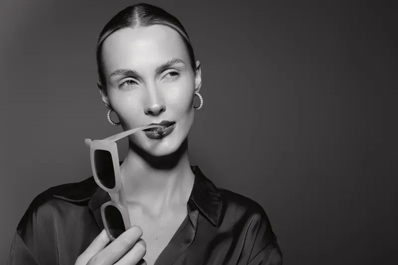 Portrait of a fair skinned white woman holding sunglasses near lips with a thoughtful expression in monochrome.