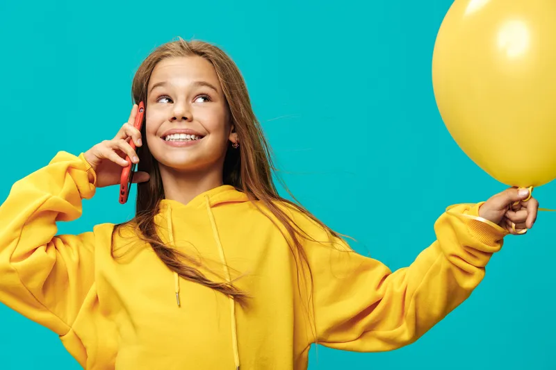 Smiling girl with yellow hoodie holding balloon and talking on phone against blue background. Happy child with long hair expressing joy and positive emotions indoors.