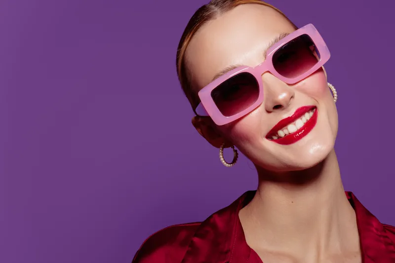 Smiling woman with red lipstick wearing pink sunglasses and hoop earrings poses against purple background. Fashion portrait with bright colors and confident expression.