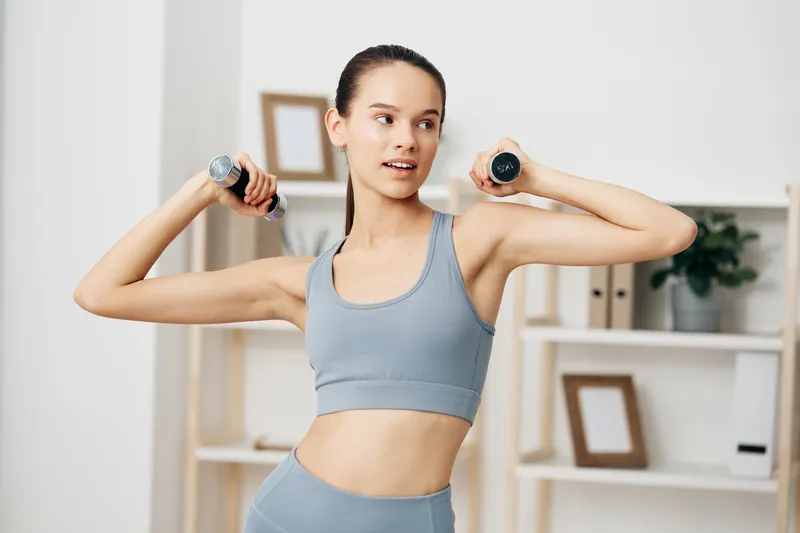 Fitness woman in sportswear lifts dumbbells in a bright home gym, showcasing exercise technique, balance and focus during a dedicated workout session for health and energy.