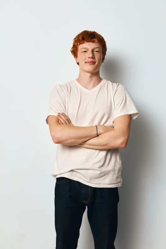 Young man with red hair standing with crossed arms in casual white t shirt and dark pants against white background with confident and relaxed expression.