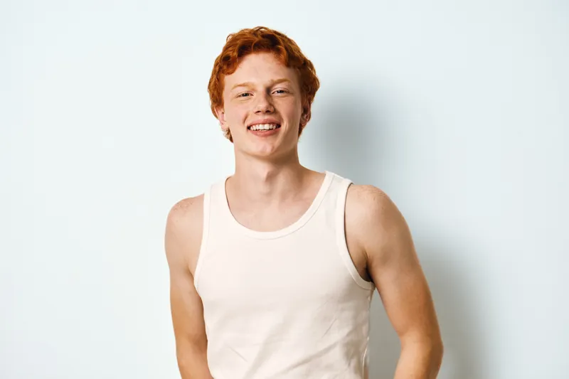 Young man with red hair and fair skin smiling in white tank top against plain white background. Casual style, happy expression and relaxed posture for lifestyle portrait.