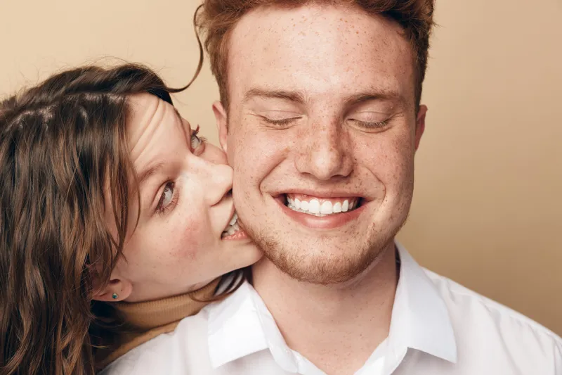 Happy young couple with freckles and casual clothes share intimate moment, woman whispering in man’s ear. Concept of love, affection, friendship, and joyful relationship in studio setting.
