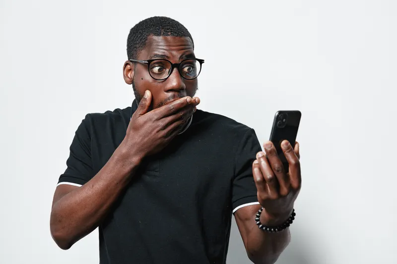 African American man with dark skin and glasses looking surprised at smartphone screen, wearing black t shirt and bracelet on white background.