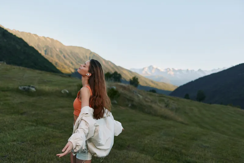 A joyful woman stands in a lush meadow with distant mountains, wearing an orange top and a light jacket, arms open, smiling, and enjoying fresh air in a scenic landscape.