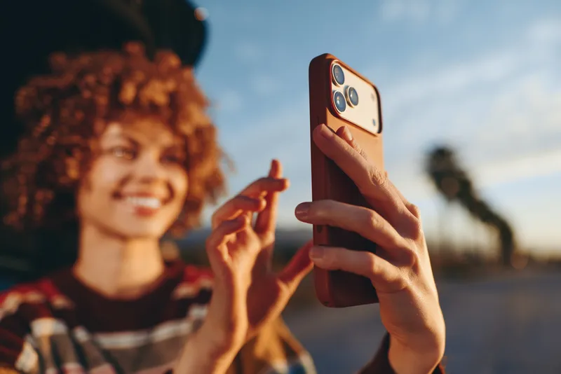 Woman lifestyle outdoor moment, smiling woman holds phone for selfie, close foreground hand and smartphone, sunset sky glow, friend blurred in background, relaxed cheerful mood, colors