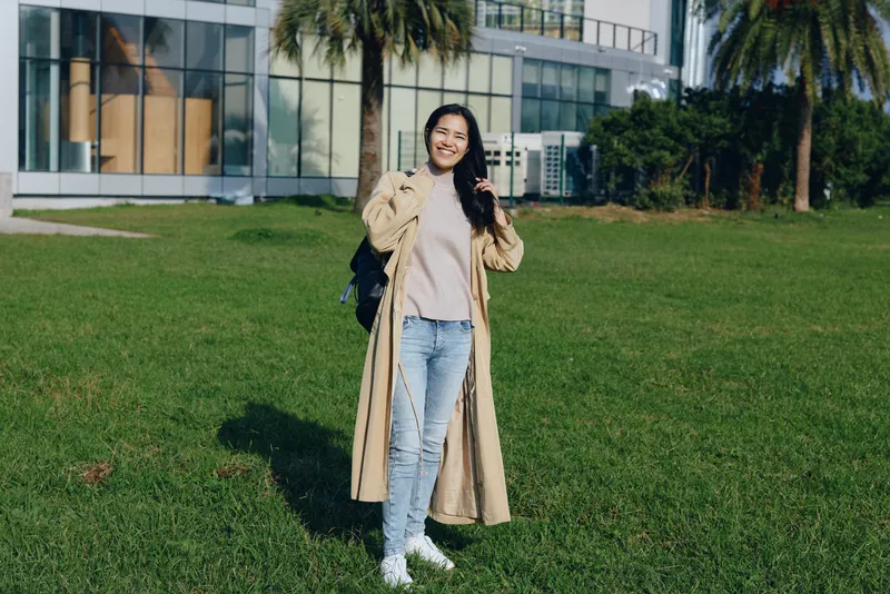 Woman in beige coat standing on a green lawn beside a glass building, casual outdoor fashion with a bag, smiling and relaxed in bright daylight, urban setting with modern architecture