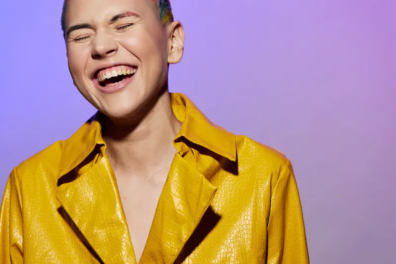 Laughing woman with short hair wearing yellow textured jacket, expressing joy and positivity against vibrant purple background, bright colorful portrait, cheerful mood.