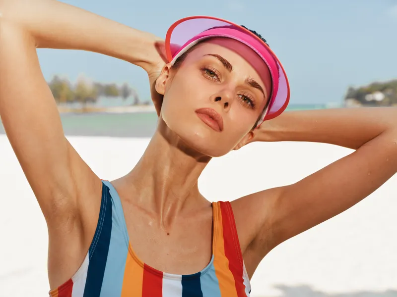 A confident female model poses on a sunlit beach, wearing a colorful striped swimsuit and pink visor, with bright sea, soft sand, and clear sky forming a vibrant scene.