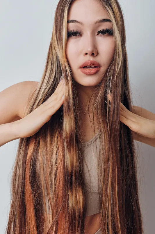 Young woman with long straight hair touching it gently standing against white background, natural beauty, hairstyle care and skin with casual wear and relaxed pose.