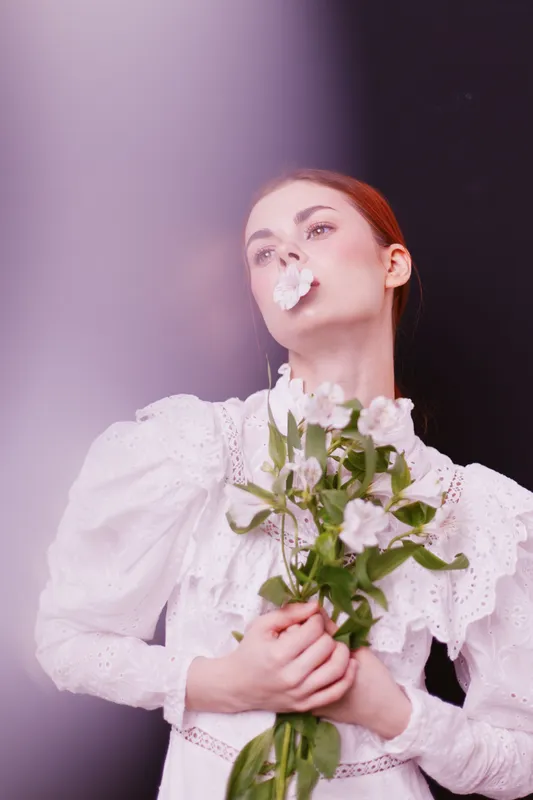 Elegant young model in vintage white dress holding flowers with a dreamy expression, showcasing beauty and femininity against a soft gradient background