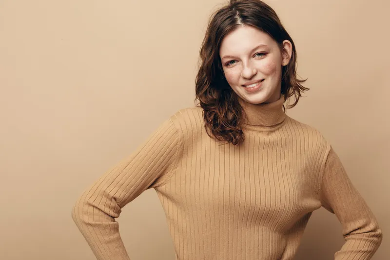Smiling young woman with brown hair wearing beige knitted sweater stands against beige background. Casual style, confident pose, natural light, portrait of happy female model.