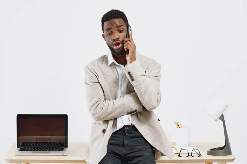 Man talking on mobile phone while standing at desk with laptop and desk lamp in office setting. Casual business environment, professional communication, technology connection, working remotely, modern