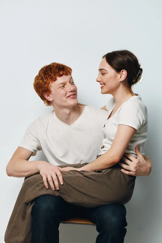 Young couple sitting close together smiling indoors studio with white background. Casual clothing, happy expressions, affectionate pose showing connection and warmth between man and woman.