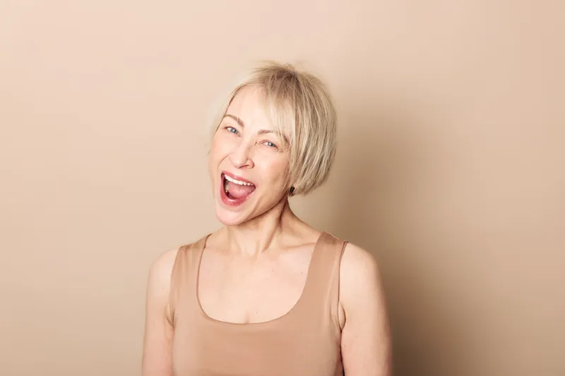 Happy middle aged woman with short blonde hair smiles and winks wearing a beige sleeveless top on neutral background. Concept of joy, confidence, natural beauty and positive emotions.
