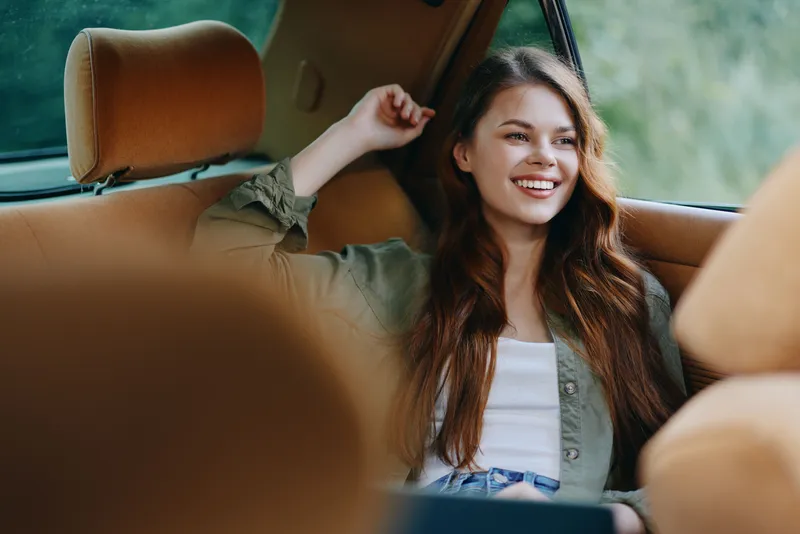 A joyful young woman with long hair sitting in a vintage car, wearing a casual outfit, exuding happiness and relaxation in a warm, natural setting.