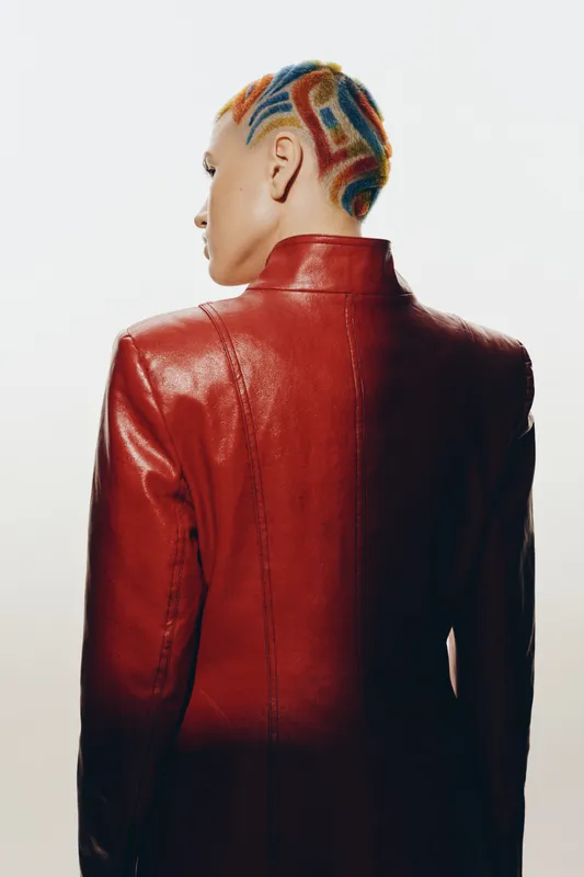 Woman with colorful patterned shaved hairstyle wearing a red leather jacket, standing with back turned against white background showcasing bold urban fashion style.