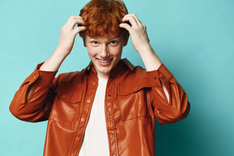 Young man with red hair wearing brown leather jacket smiling and touching hair against turquoise background in studio. Casual style, confident mood and youth fashion portrait.