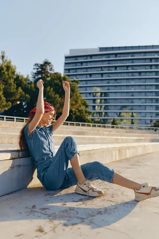 young woman sitting outdoors in casual denim outfit raising hands with joyful expression under clear sky in urban setting