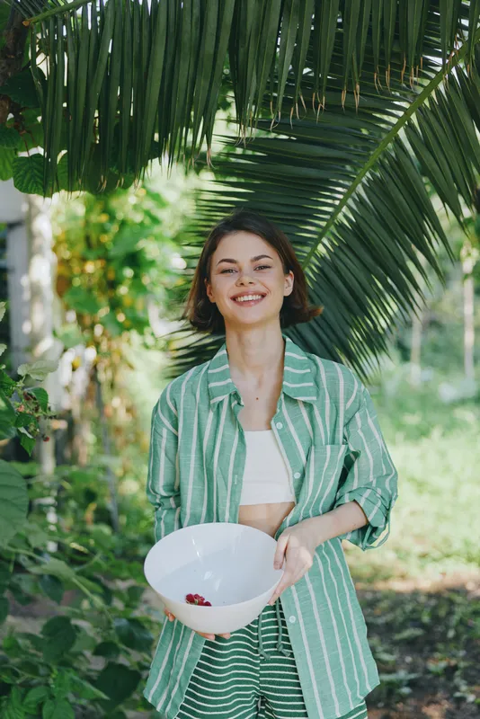 A cheerful woman stands outdoors among lush greenery, wearing a striped green shirt and light outfit, holding a white bowl with berries, radiating joy, style, and a relaxed mood in bright sunlight.