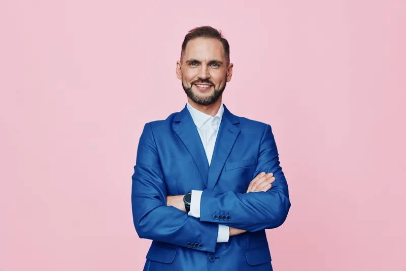 A professional man in a blue suit stands with arms crossed against a solid pink background, projecting confidence, calm focus, and leadership presence for business visuals.