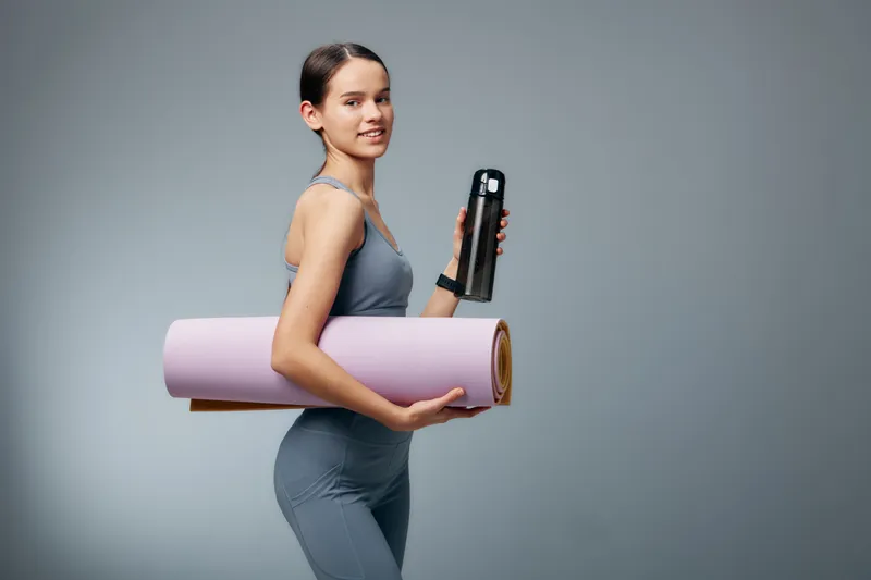 Woman in athletic wear carries a yoga mat and water bottle, ready for a workout. She stands in a bright studio, embodying fitness, sport and active training.
