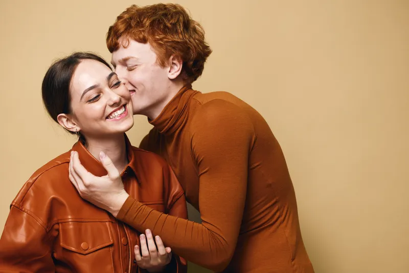 Young couple with joyful expressions hugging and kissing each other against beige background. They wear brown casual clothes showing affection and intimacy in studio portrait.