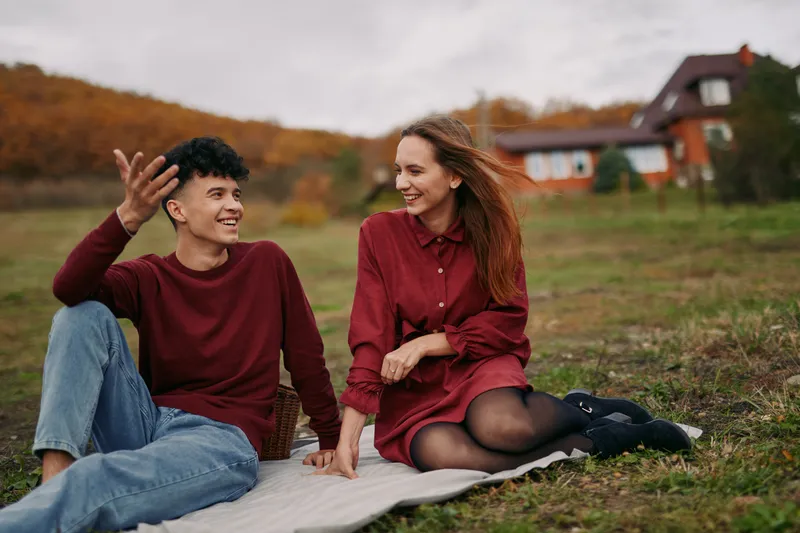 A candid outdoor moment features two friends sitting on a blanket, sharing a genuine smile and easy conversation under soft autumn light, conveying authenticity.