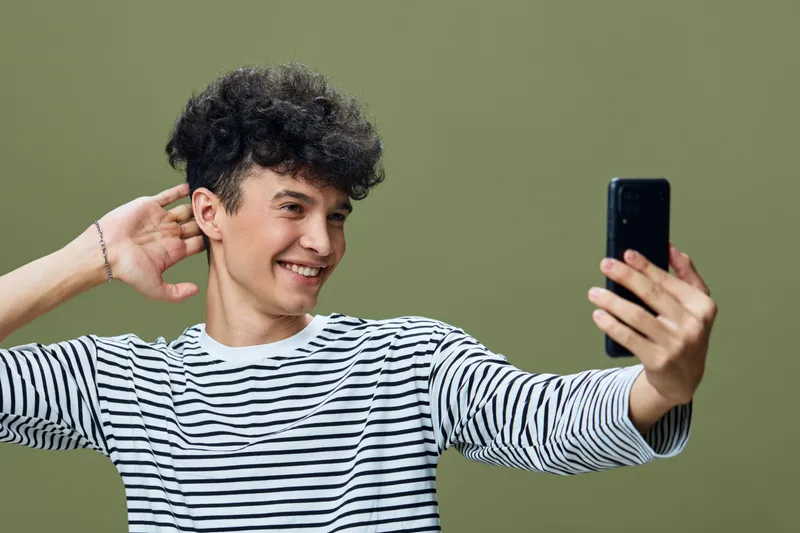Smiling young man in striped shirt taking selfie with smartphone, casual hairstyle, soft green background, cheerful expression, modern lifestyle concept.
