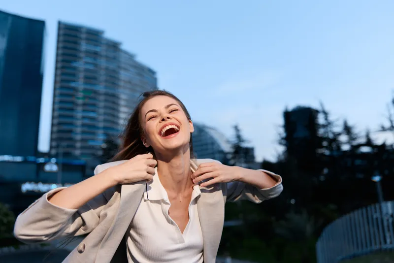 Happy woman outdoors with casual beige jacket laughing joyfully in urban environment under bright blue sky smiling cheerful emotion business concept