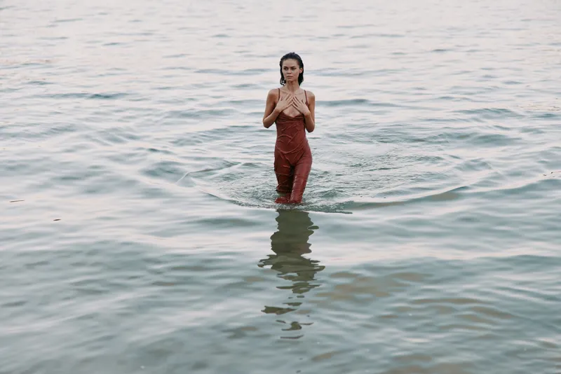 A woman stands waist-deep in calm sea water, wearing a red dress, creating gentle ripples and a serene, contemplative mood.