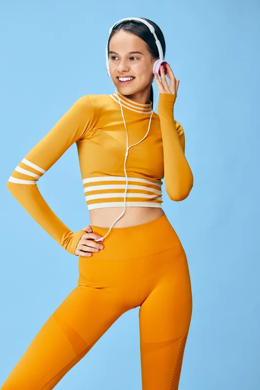 Fit young woman in orange athletic crop top and high waisted leggings wearing white headphones, posing confidently with hand on hip in a bright blue studio, sporty lifestyle concept.