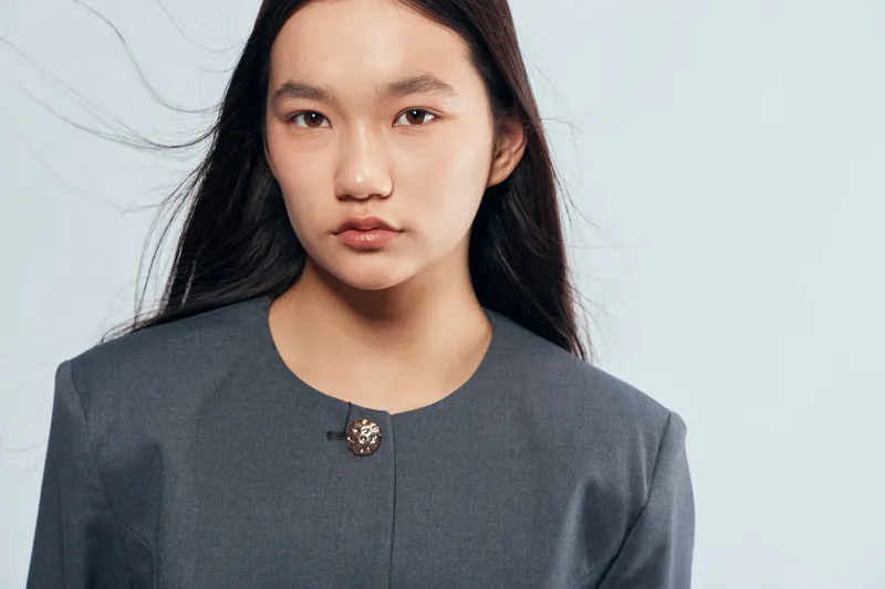 Portrait of a young woman with long dark hair in a grey jacket looking at camera with neutral expression on light background. Closeup of female face and upper body for fashion.