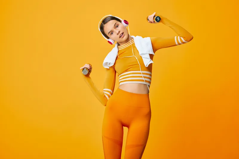 Fitness, woman in bright orange athletic wear poses mid workout with dumbbells, headphones and a towel draped over shoulders against a bold orange backdrop.