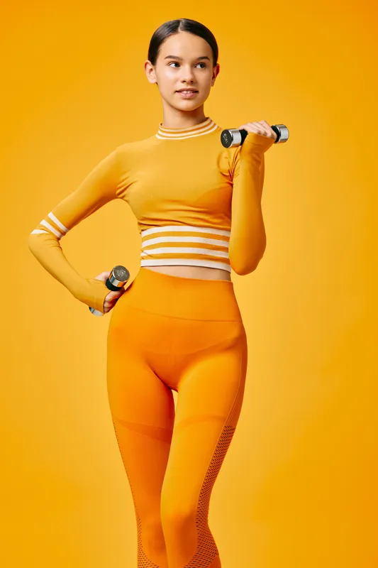 Fitness woman in orange athletic wear lifts dumbbells in a vibrant orange studio, showcasing strength, balance, and confidence during a focused workout session.